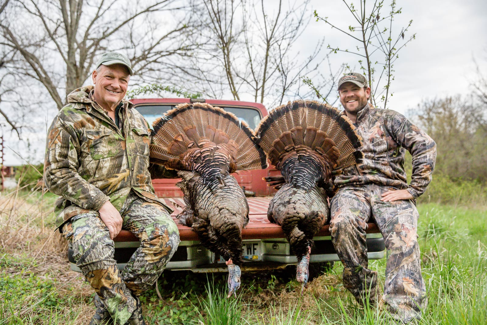Two hunters in camouflage sit on the tailgate of a red truck with two harvested wild turkeys.