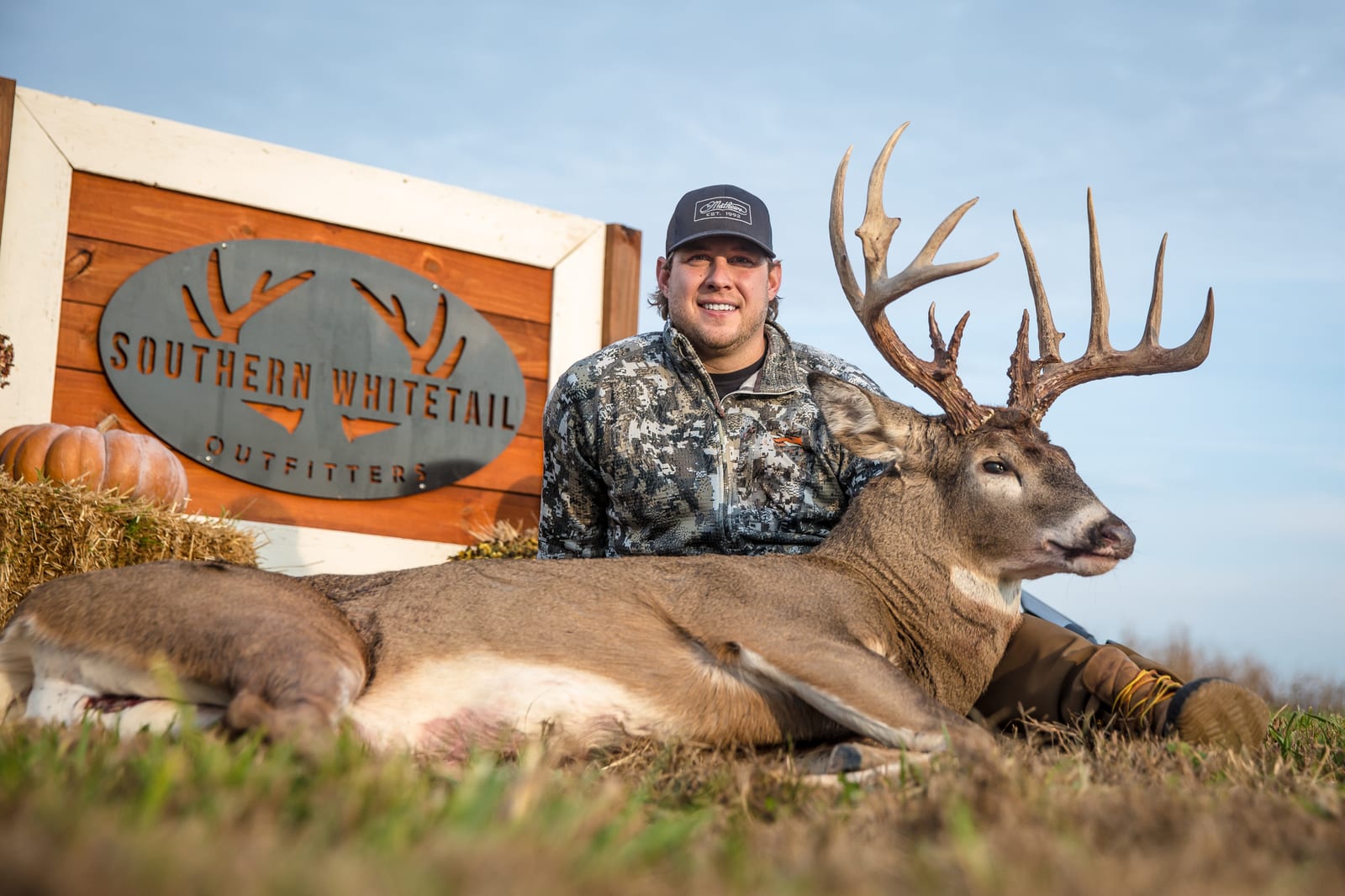 A hunter poses with a harvested whitetail deer in front of a "Southern Whitetail Outfitters" sign.