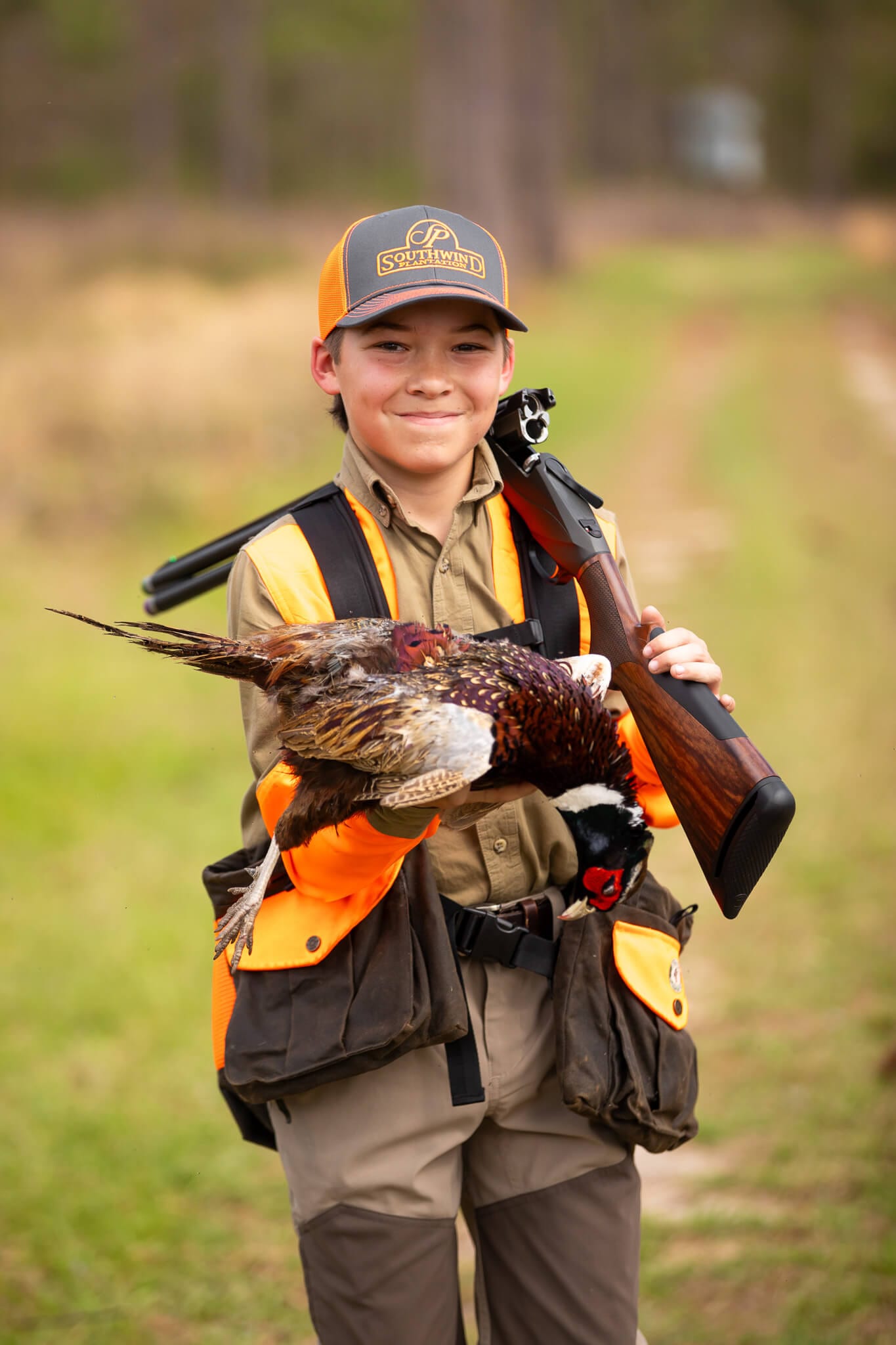A young hunter holds a harvested ring-necked pheasant and a shotgun.