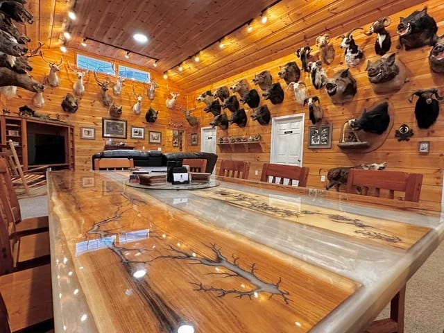 The interior of a hunting lodge features wooden walls adorned with numerous taxidermy animal mounts, a large epoxy river table, and a seating area with a television.