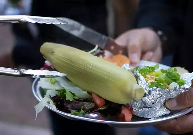 A person serves a plate with corn on the cob, a baked potato, and a salad.