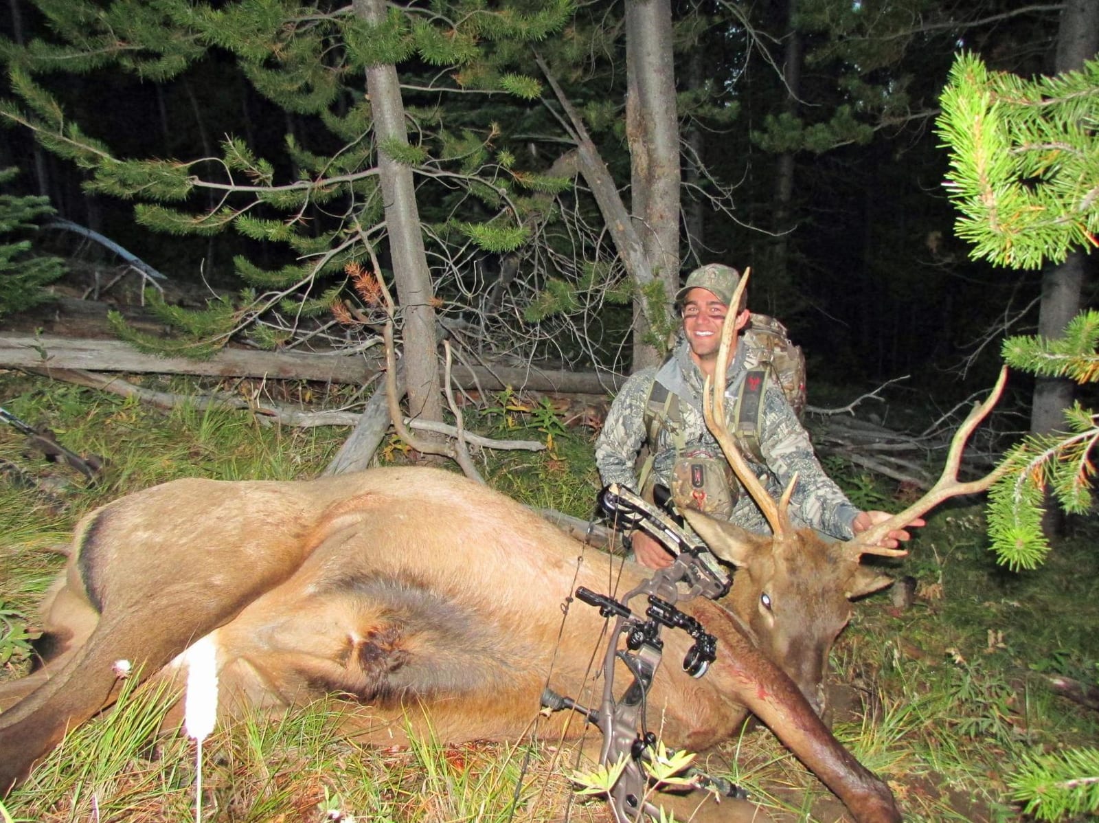 A hunter in camouflage poses with a harvested elk in a forest setting.