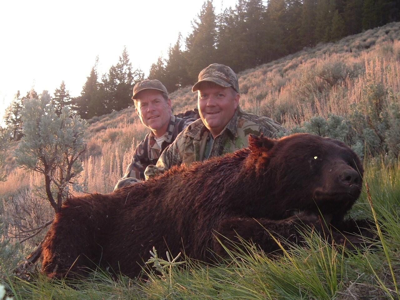 Two hunters pose with a harvested black bear on a grassy hillside.