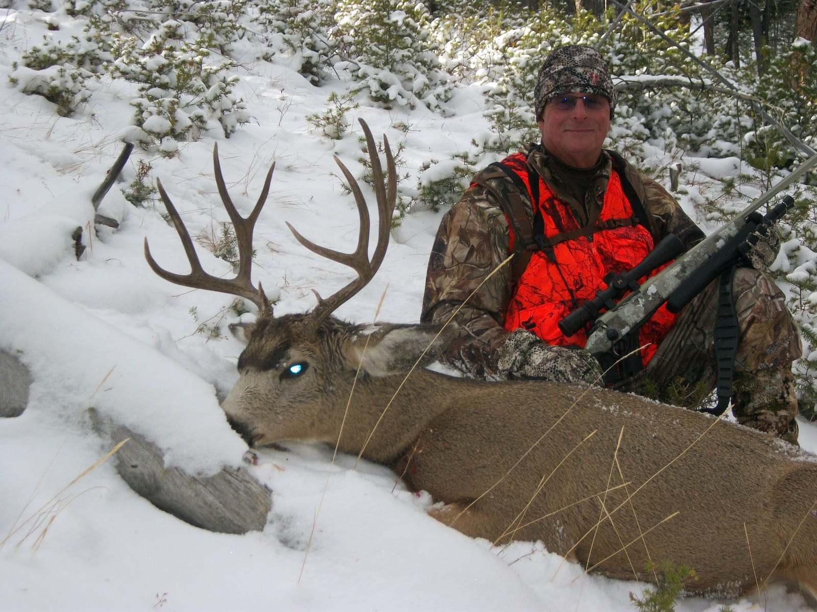 A hunter poses with a harvested mule deer in a snowy landscape.