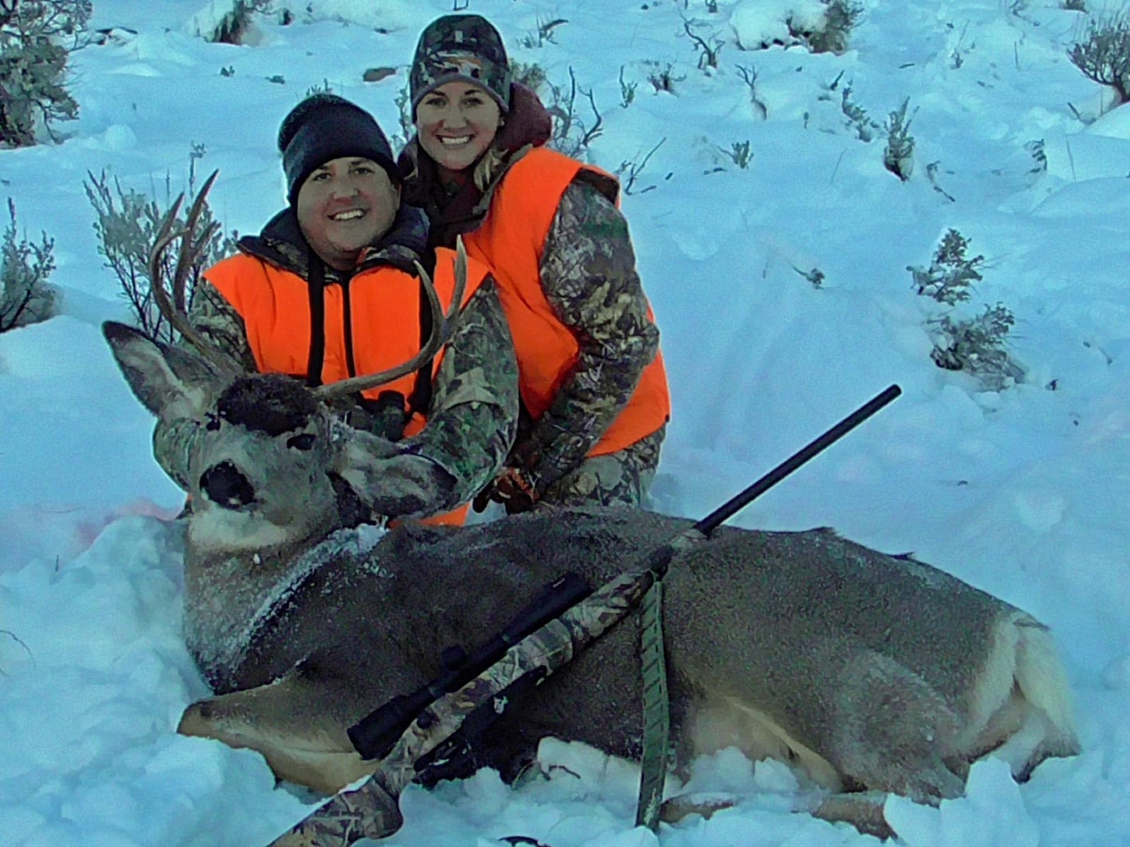 Two hunters in orange vests pose with a harvested mule deer in a snowy landscape.