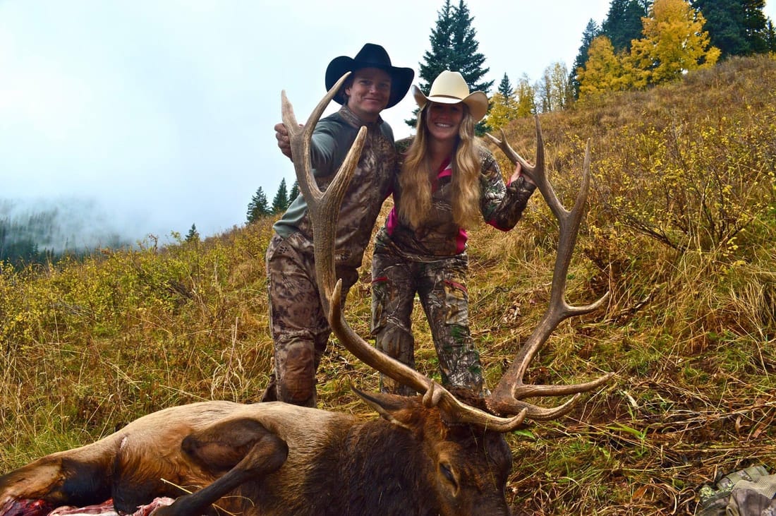Two hunters pose with a harvested elk in a grassy, mountainous field.