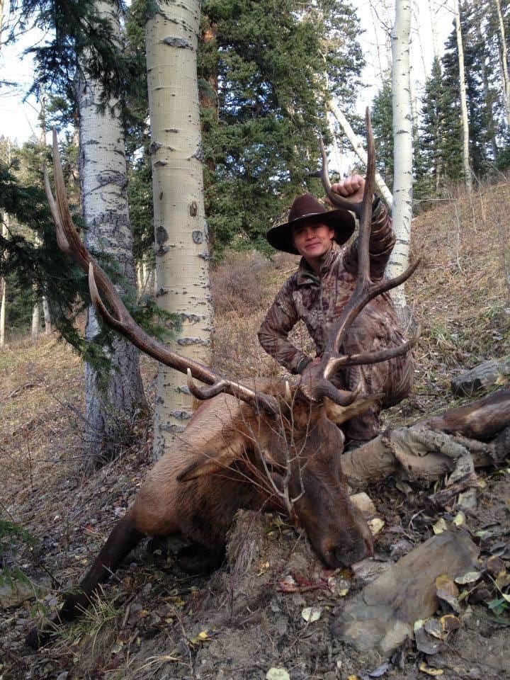A hunter in camouflage and a hat poses with a harvested elk in a wooded area.