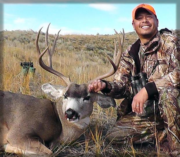 A hunter in camouflage and an orange hat poses with a harvested mule deer in a grassy field.