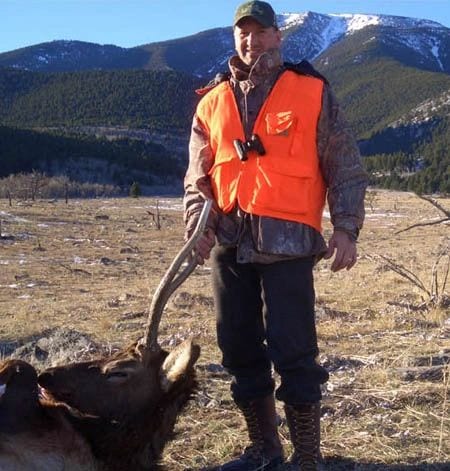 A hunter in an orange vest poses with a harvested elk in a field with mountains in the background.