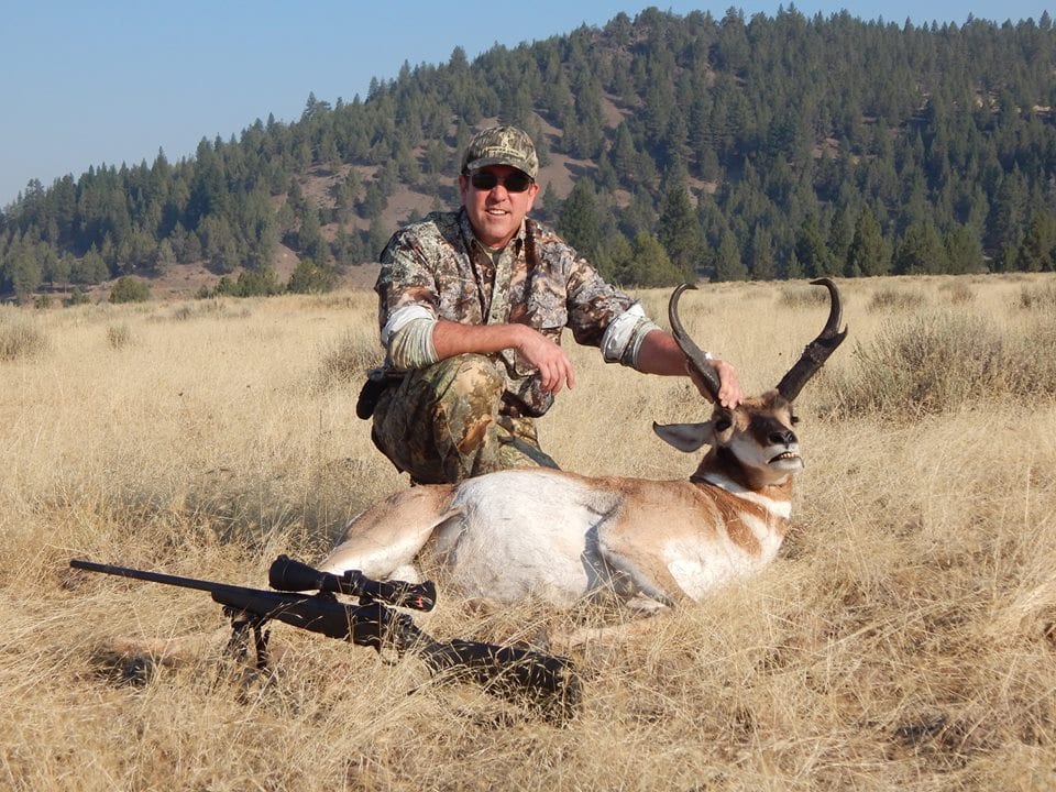 A hunter kneels next to a harvested pronghorn in a grassy field with a rifle on the ground.