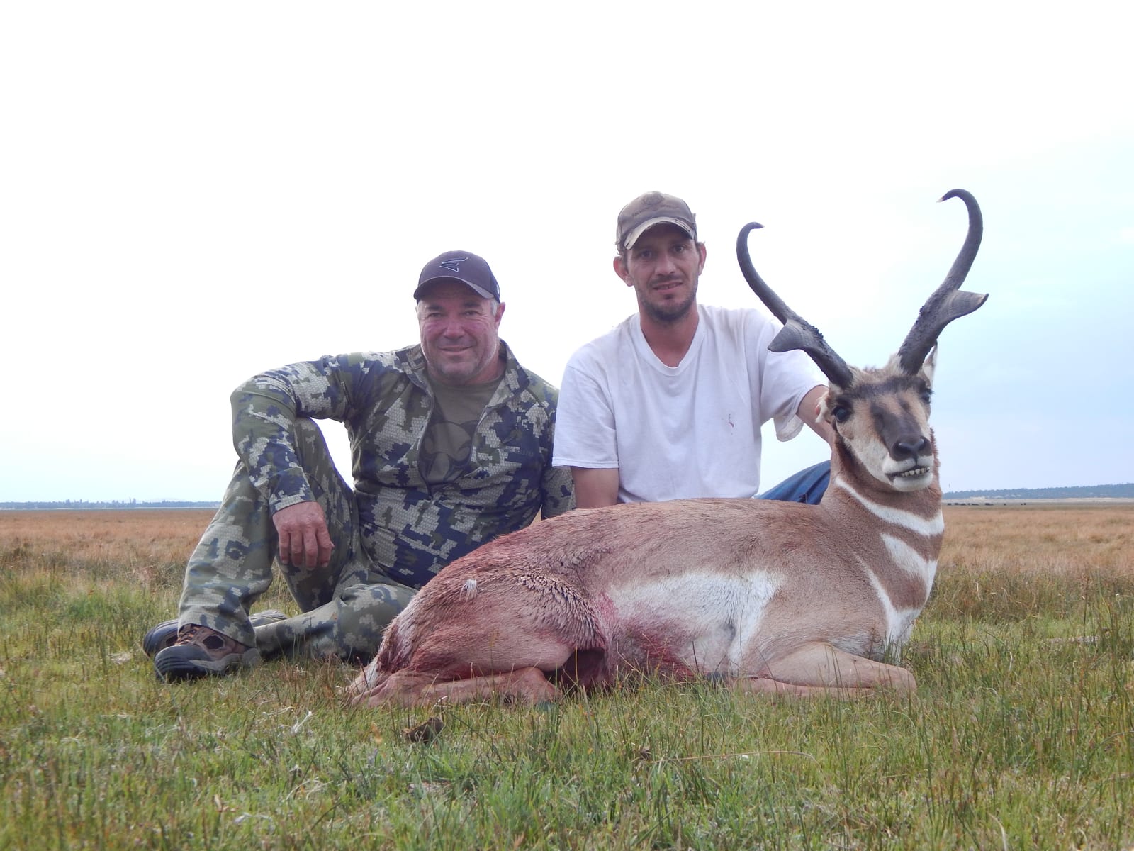 Two hunters pose with a harvested pronghorn in a grassy field.