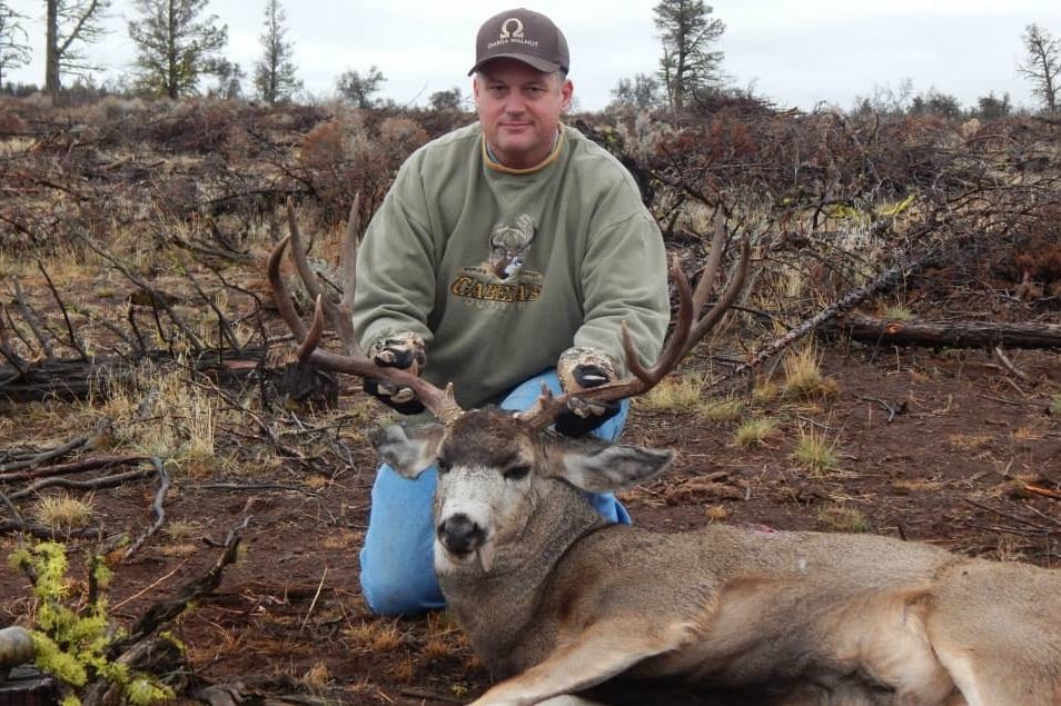 A hunter kneels with a harvested mule deer in a dry, brushy landscape.