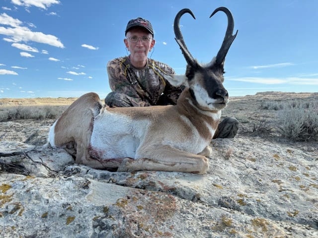 A hunter poses with a harvested pronghorn in a field under a blue sky.