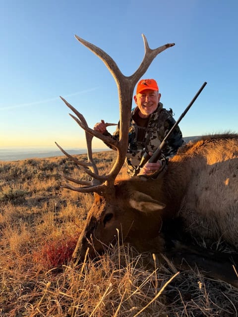 A hunter in an orange hat poses with a harvested elk on a grassy hillside.
