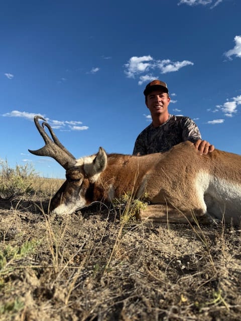 A hunter poses with a harvested pronghorn under a blue sky.