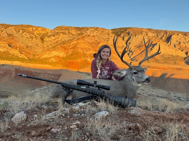 A hunter poses with a harvested mule deer in a mountainous landscape.