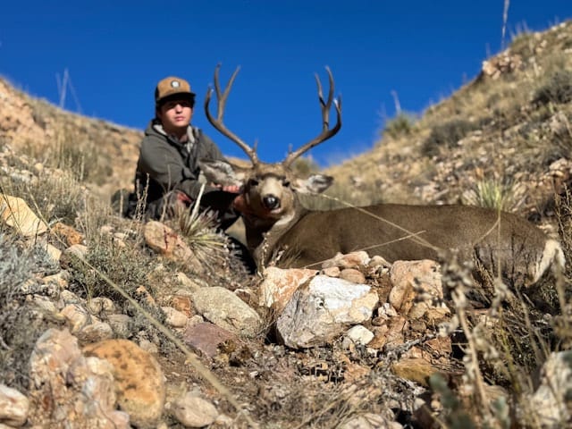 A hunter poses with a harvested mule deer on a rocky, grassy hillside under a clear blue sky.