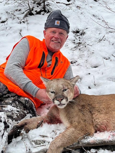 A man in an orange vest poses with a harvested mountain lion in the snow.