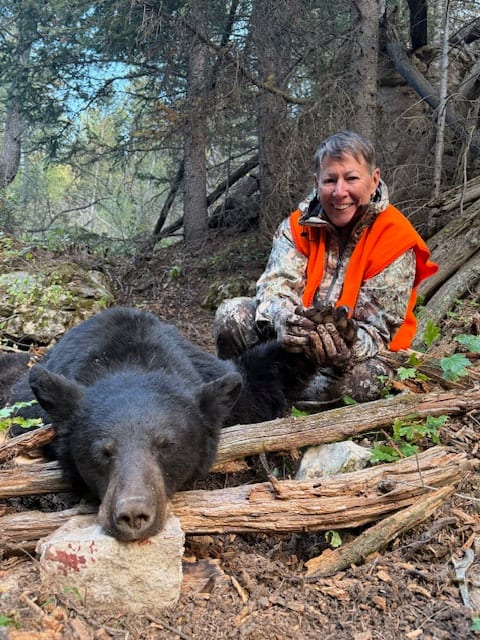 A woman in hunting gear poses with a harvested black bear in a forest.