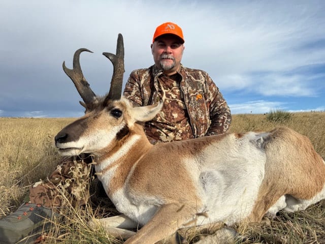 A hunter in camouflage and an orange hat poses with a harvested pronghorn in a grassy field under a cloudy sky.