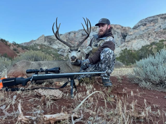 A hunter in camouflage poses with a harvested mule deer and a rifle in a rocky, arid landscape.