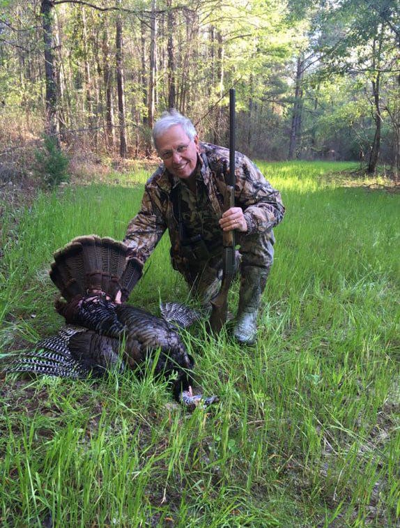 A hunter in camouflage kneels next to a harvested wild turkey in a grassy field with trees in the background.