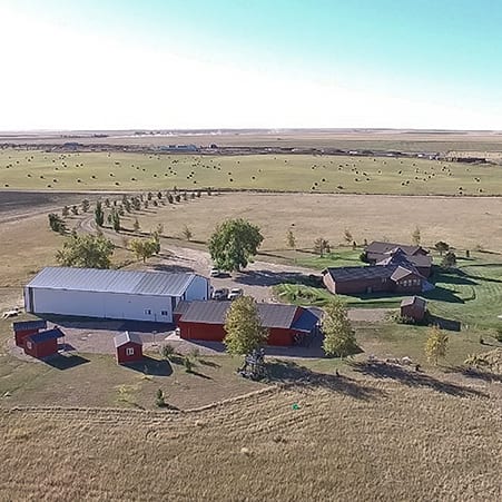 An aerial view of a large property featuring multiple buildings, including a main lodge-style structure and several barns, surrounded by expansive fields with hay bales.