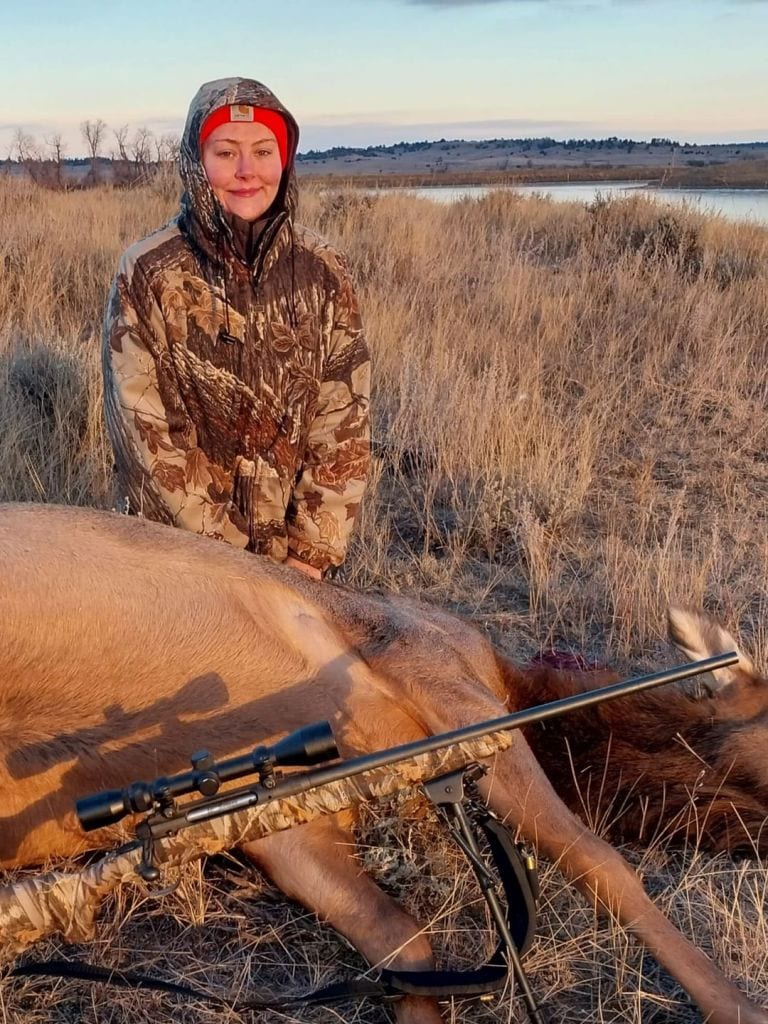 A woman in camouflage and a red hat poses with a harvested elk and a rifle in a grassy field with a body of water in the background.