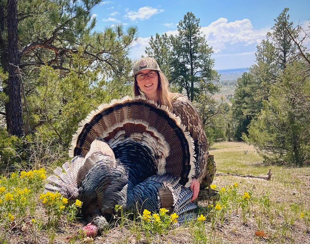 A woman poses with a harvested wild turkey in a natural setting.