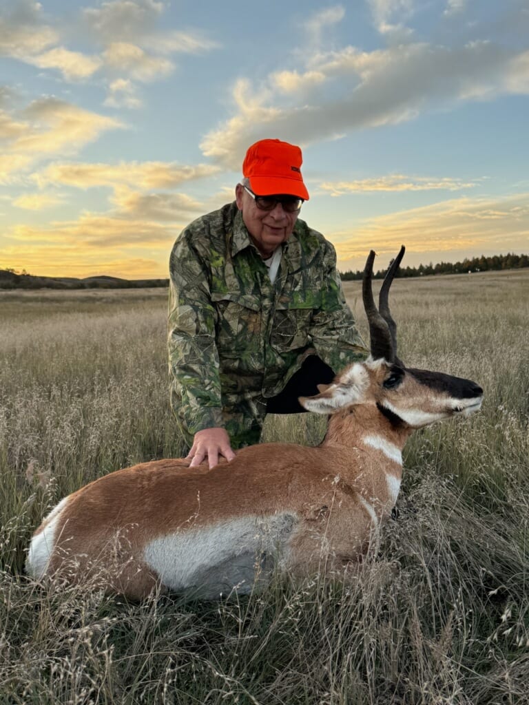 A hunter in camouflage and an orange hat poses with a harvested pronghorn in a grassy field at sunset.