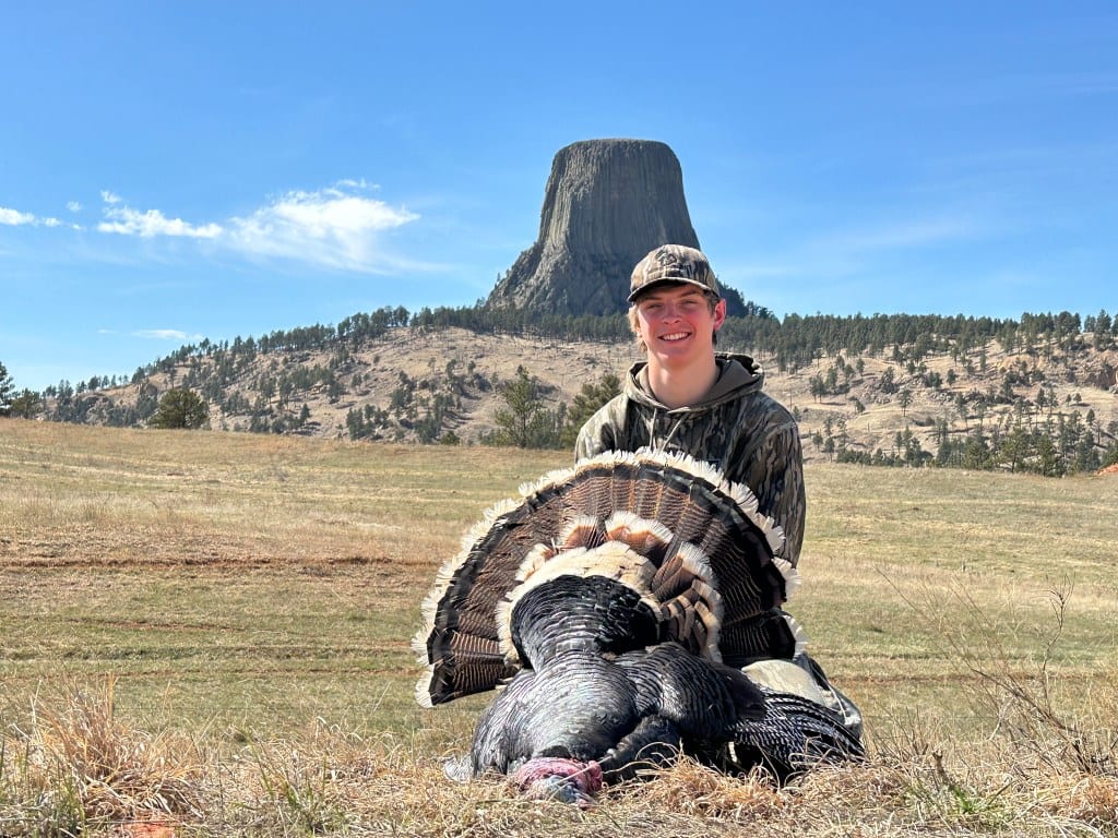 A hunter poses with a harvested wild turkey in a field with Devils Tower in the background.