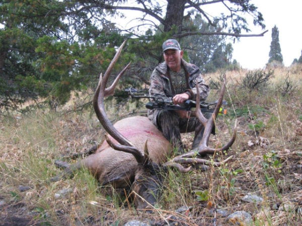 A hunter in camouflage sits next to a harvested elk with large antlers, holding a bow.