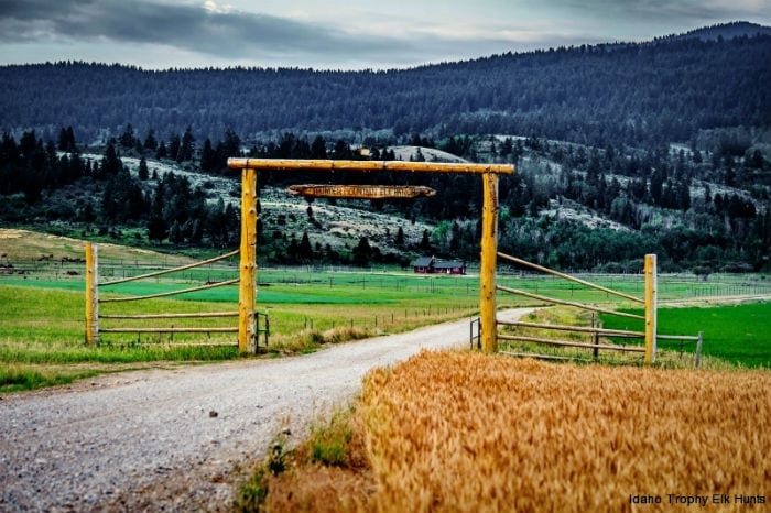 A dirt road leads to a wooden gate with a sign, flanked by fields and forested mountains under a cloudy sky.
