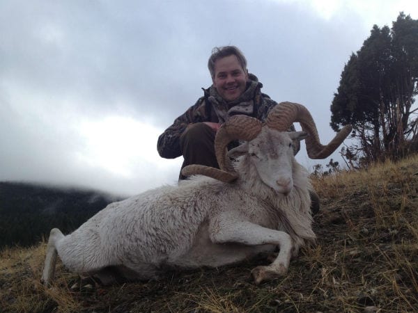 A hunter poses with a harvested Dall sheep on a grassy hillside under an overcast sky.