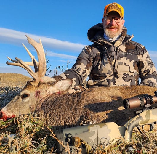 A man in camouflage poses with a harvested whitetail deer and a rifle.