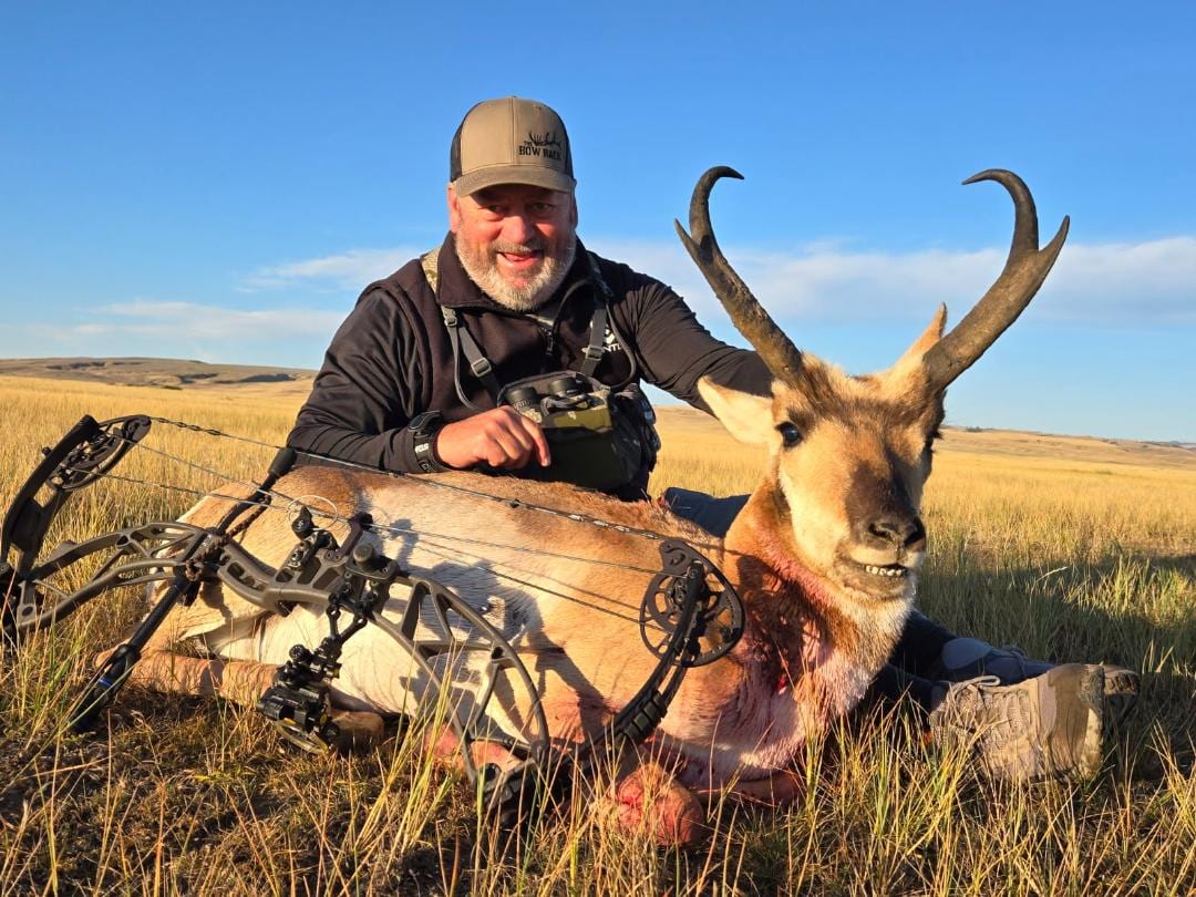 A man poses with a harvested pronghorn and a bow in a grassy field under a blue sky.