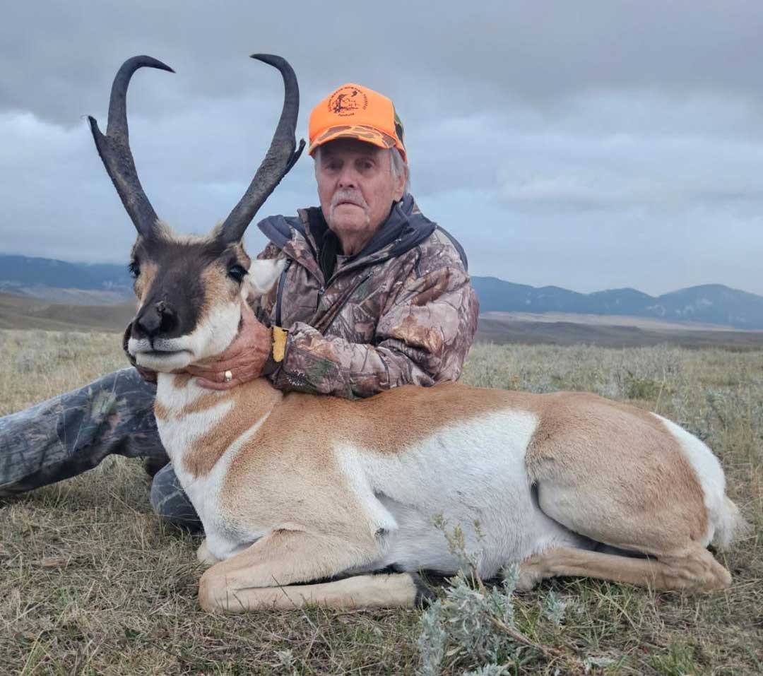 An older man in camouflage and an orange hat poses with a harvested pronghorn in a grassy field.