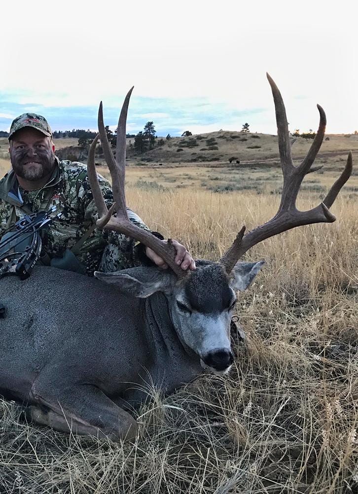 A hunter in camouflage poses with a harvested mule deer in a grassy field.