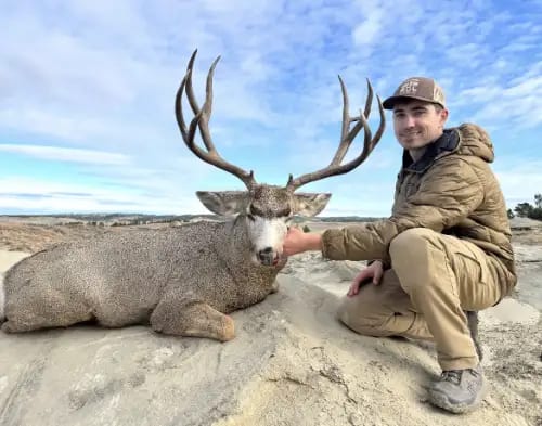 A hunter poses with a harvested mule deer in a field.
