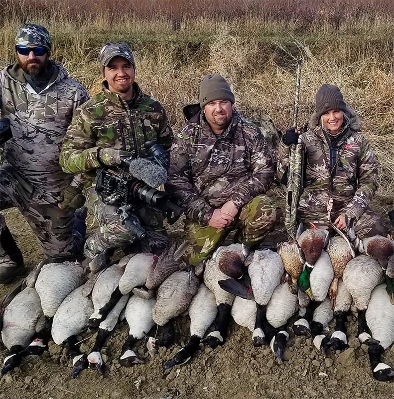 Four hunters pose with their harvest of Canada geese and pheasants in a field.