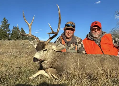 Two hunters pose with a harvested mule deer in a grassy field under a blue sky.