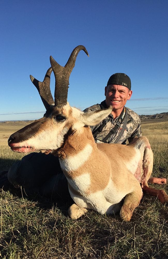 A hunter smiles while posing with a harvested pronghorn in a grassy field under a clear blue sky.
