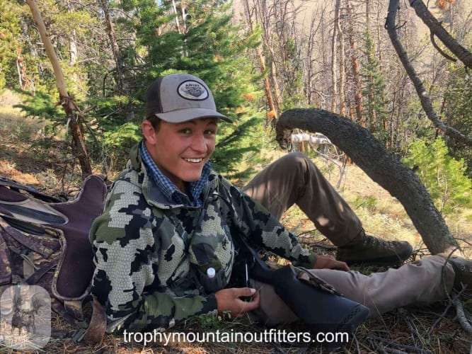 A hunter in camouflage and a hat is sitting on the ground in a forest, holding a rifle and smiling.