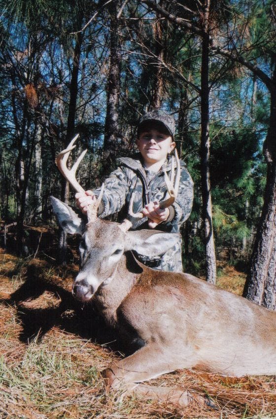 A young hunter poses with a harvested whitetail deer in a forest.