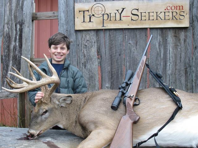 A young hunter poses with a harvested whitetail deer and a rifle.