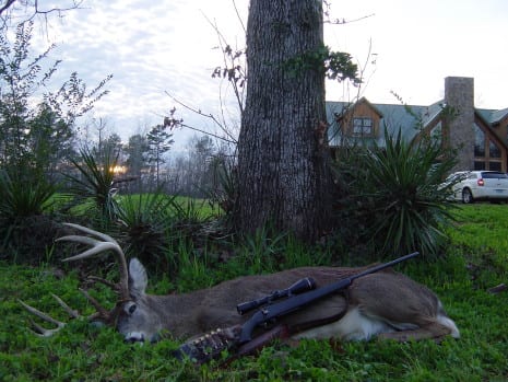 A harvested whitetail deer lies in the grass with a rifle next to it, with a house in the background.