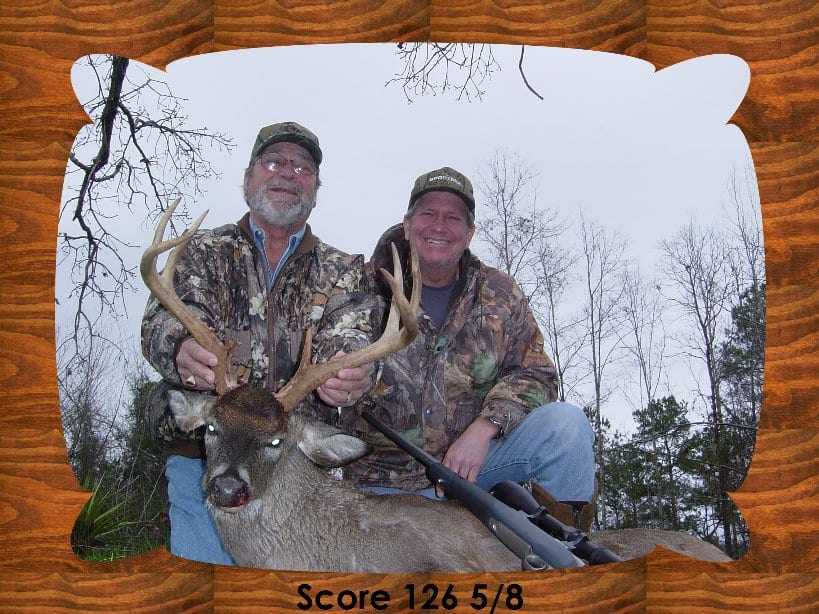 Two hunters pose with a harvested whitetail deer in a wooded area, framed by a wooden border.