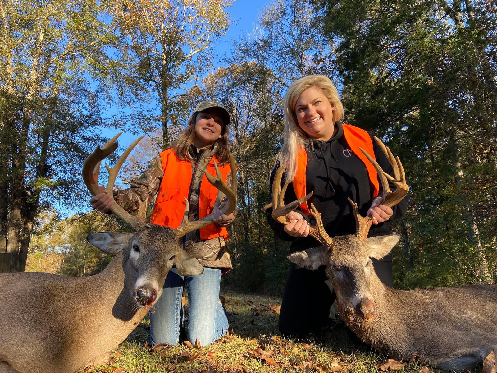 Two women in orange vests pose with two harvested whitetail deer in a wooded area.
