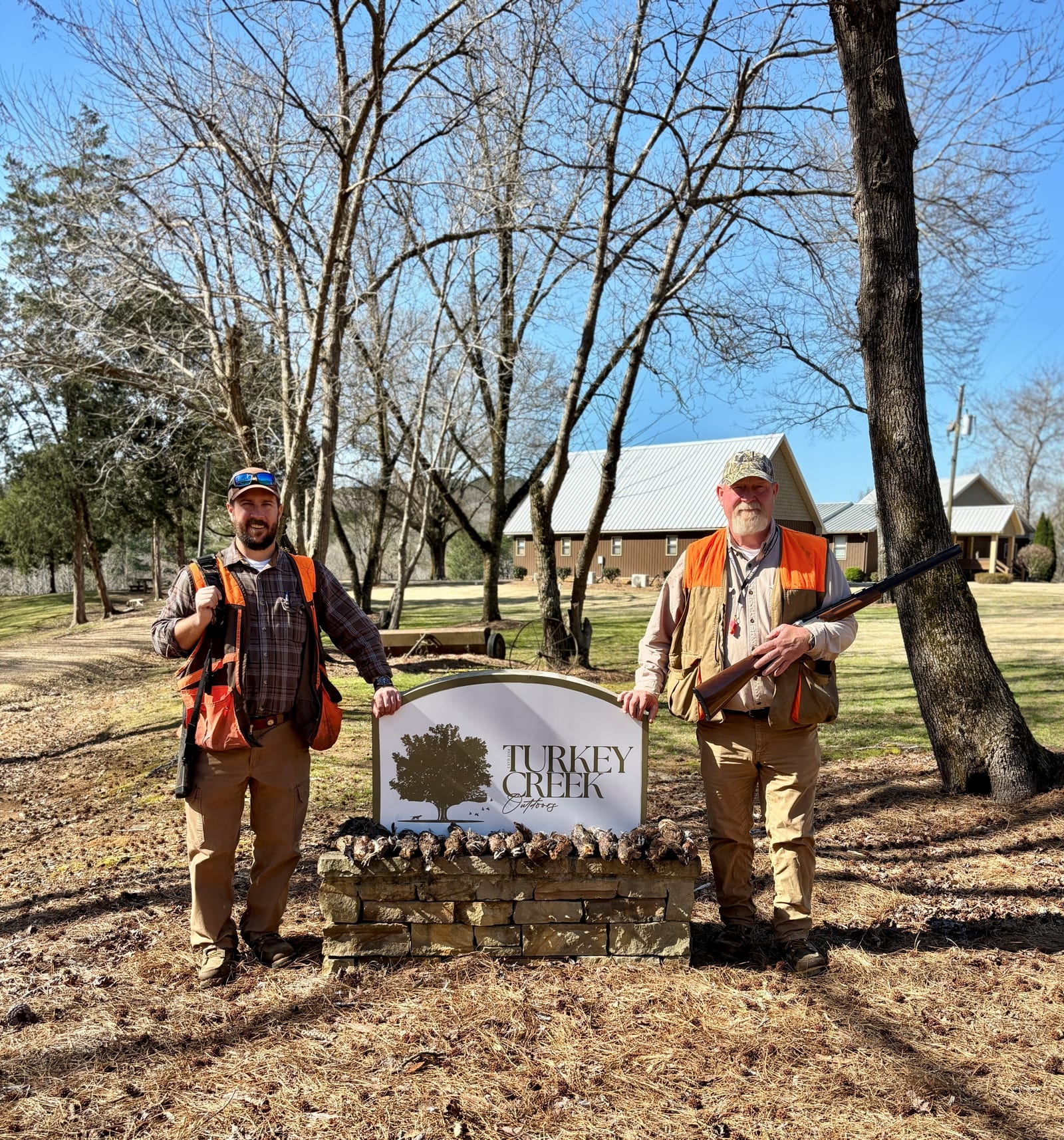 Two men in hunting vests stand in front of a "Turkey Creek" sign, one holding a shotgun.