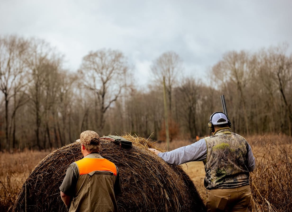 Two hunters are in a field with a hay bale and trees in the background.